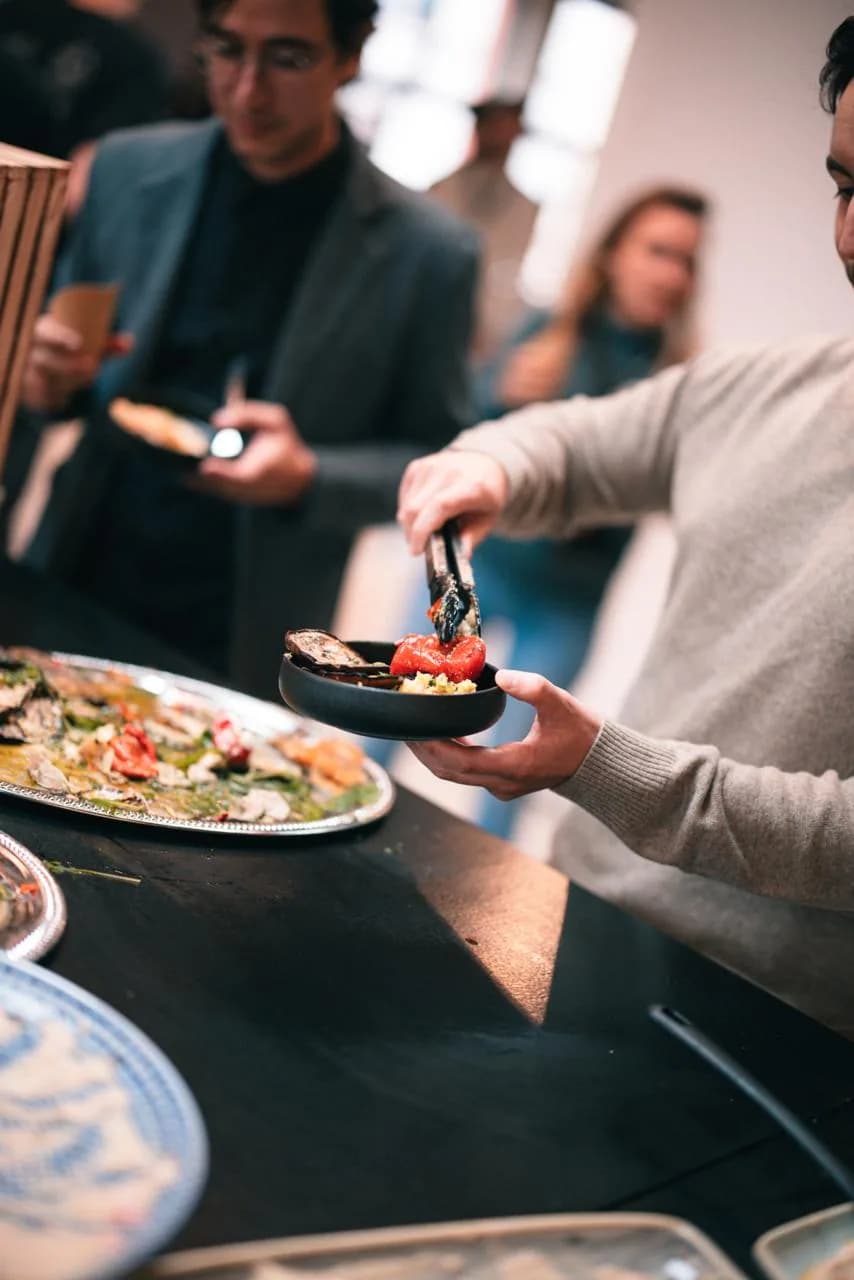 Ein Gast legt sich mit der Zange gegrilltes Gemüse und Couscous in eine Schale, dahinter weitere Gäste