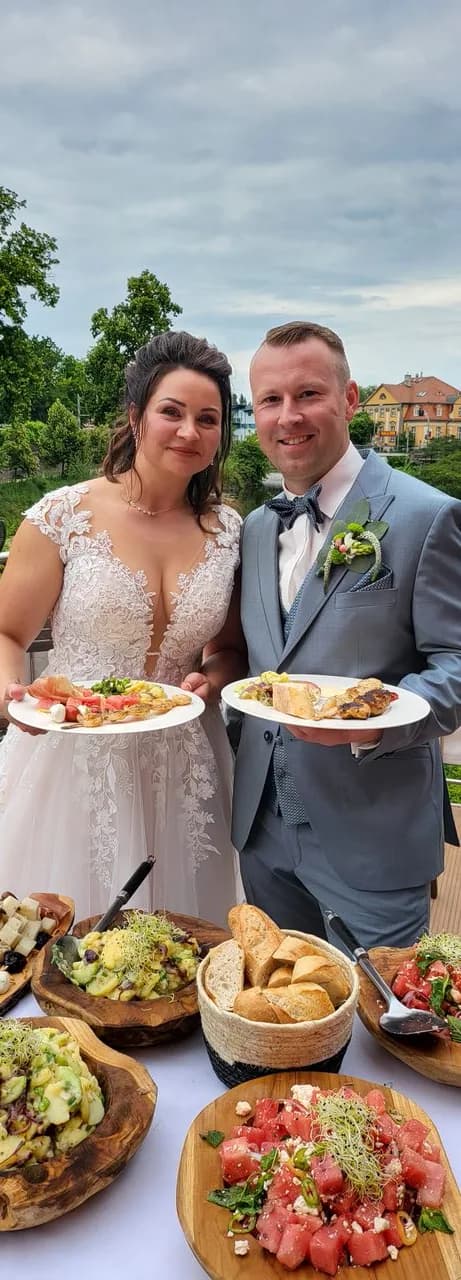 A bride and groom holding plates of starters, with a buffet of salads and bread in front